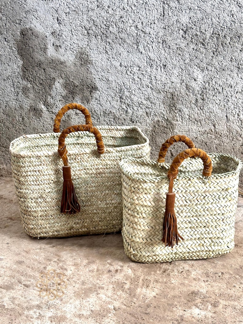 May include: Two rectangular woven straw baskets with brown leather handles and tassel details. The baskets are of different sizes and have a natural colour. They are set against a textured grey background.
