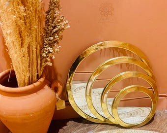 Könnte beinhalten: Ein Satz von vier goldfarbenen, kreisförmigen Spiegeln in verschiedenen Größen. Die Spiegel sind in einem verschachtelten Muster angeordnet. Links befindet sich eine Terrakotta-Vase mit getrockneten Pflanzen. Die Spiegel stehen auf einer gewebten Matte.