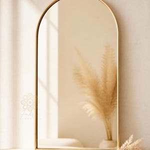 K&ouml;nnte beinhalten: Ein bogenf&ouml;rmiger Wandspiegel mit einem d&uuml;nnen, polierten Goldmetallrahmen. Die Spiegelfl&auml;che reflektiert einen neutralen Innenraum mit einer Vase, die getrocknetes Pampasgras enth&auml;lt. Ein kleines Logo ist unten links zu sehen.