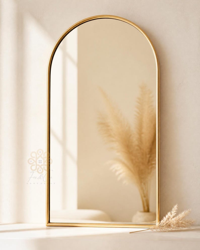 K&ouml;nnte beinhalten: Ein bogenf&ouml;rmiger Wandspiegel mit einem d&uuml;nnen, polierten Goldmetallrahmen. Die Spiegelfl&auml;che reflektiert einen neutralen Innenraum mit einer Vase, die getrocknetes Pampasgras enth&auml;lt. Ein kleines Logo ist unten links zu sehen.