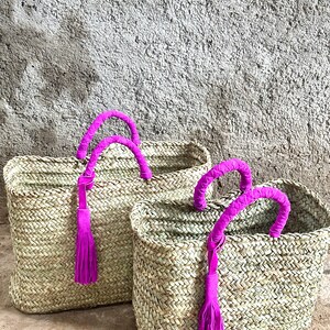 May include: Two woven straw tote bags with bright pink handles and tassels. The bags are rectangular with a basket weave pattern. The handles are wrapped in pink fabric, and the tassels hang down from the handles.