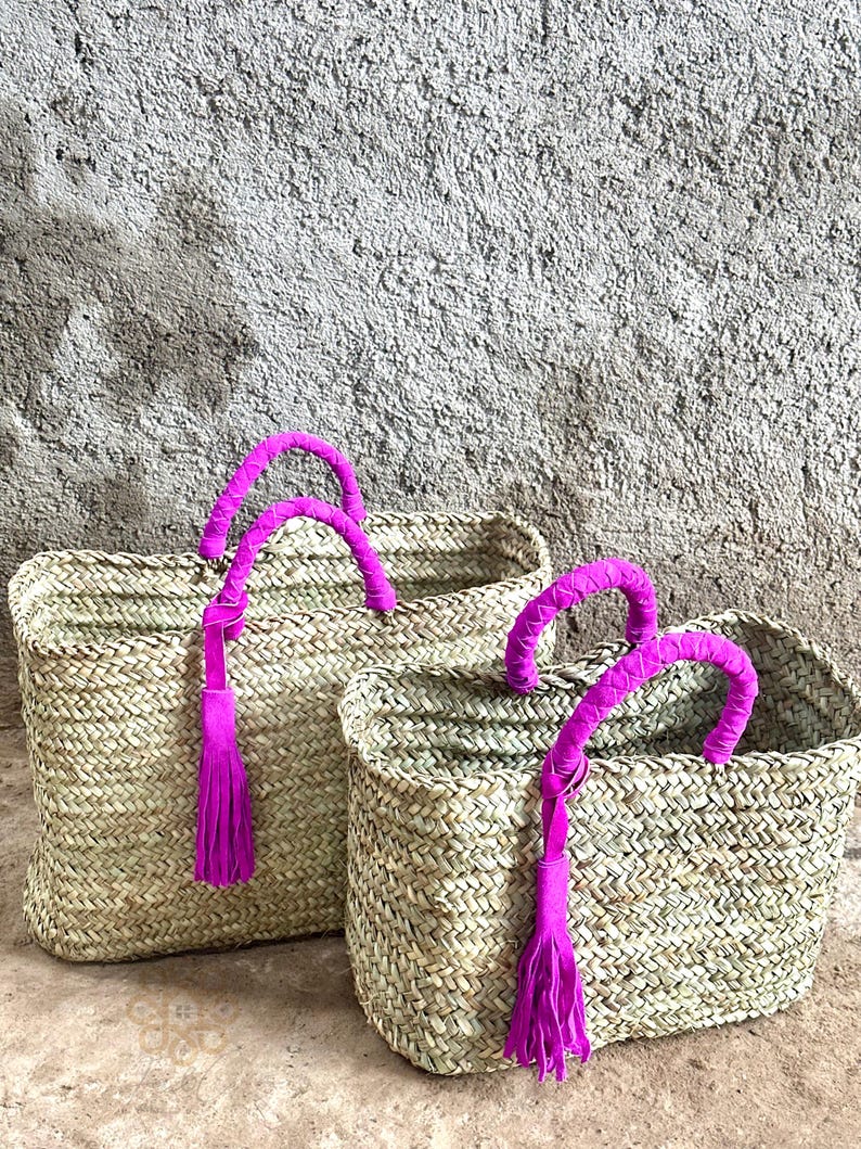 May include: Two woven straw tote bags with bright pink handles and tassels. The bags are rectangular with a basket weave pattern. The handles are wrapped in pink fabric, and the tassels hang down from the handles.