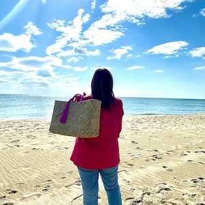 May include: A woven straw tote bag with a purple tassel and handles. The bag is carried by a person wearing a red top and blue jeans on a sandy beach with the ocean in the background. The sky is blue with white clouds.