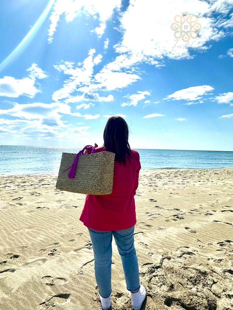 May include: A woven straw tote bag with a purple tassel and handles. The bag is carried by a person wearing a red top and blue jeans on a sandy beach with the ocean in the background. The sky is blue with white clouds.