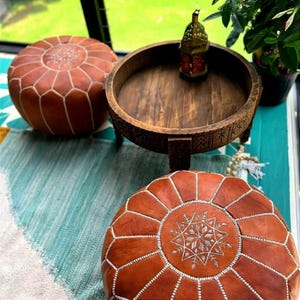 Può includere: Due pouf in pelle marrone con cuciture bianche e un tavolo in legno con gambe intagliate. I pouf hanno un motivo a stella sulla parte superiore.