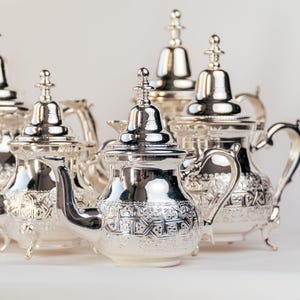 Può includere: Un set di cinque teiere d'argento con intricati disegni in rilievo. Le teiere hanno uno stile marocchino tradizionale e sono realizzate in metallo lucido.