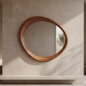 May include: A wall-mounted mirror featuring an irregular, organic, pebble-shaped wooden frame with a smooth, warm brown finish. The reflective glass surface shows a partial view of a green plant reflected within the mirror's centre.