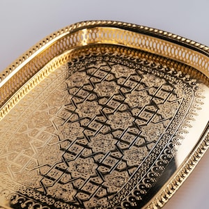 Può includere: Un vassoio da portata dorato con un intricato motivo geometrico in rilievo sulla superficie. Il vassoio ha un bordo decorativo con un design traforato.