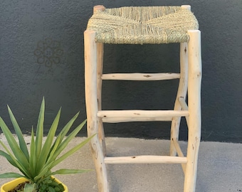 May include: A handcrafted wooden stool featuring a woven seat. The stool has four pale wooden legs and three horizontal supports. The seat is made of woven natural fibres. A yellow potted plant with green leaves is in the foreground.