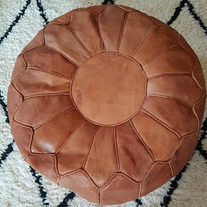 May include: A brown leather pouf with a circular top and a stitched pattern. The pouf is made of multiple panels of leather that are stitched together to create a unique design.