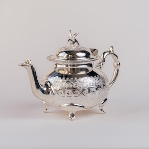 Könnte beinhalten: Eine silberne Teekanne mit einem verzierten Design und einem Hasen-Knauf auf dem Deckel. Die Teekanne hat drei Beine und einen Ausguss.