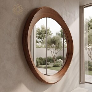 May include: A circular wall mirror featuring a thick, polished dark brown wooden frame with a smooth, curved profile. The frame has a distinct, asymmetrical depth that tapers around the circumference, reflecting a garden view with trees and shrubs.