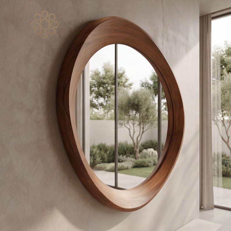 May include: A circular wall mirror featuring a thick, polished dark brown wooden frame with a smooth, curved profile. The frame has a distinct, asymmetrical depth that tapers around the circumference, reflecting a garden view with trees and shrubs.