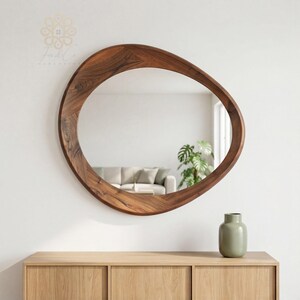 May include: A wall-mounted mirror with an irregular, organic-shaped frame crafted from dark brown wood with a visible grain pattern. The mirror reflects a room interior, and a light green ceramic vase sits on a wooden cabinet below it.