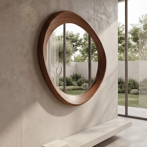 May include: A circular wall mirror featuring a thick, smooth, natural brown wood frame with an organic, asymmetrical shape. The reflective glass surface shows a clear view of an outdoor garden area with green trees, shrubs, and a lawn.