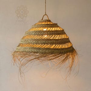 May include: A conical pendant light fitting crafted from woven natural tan fibres. The shade features a repeating braided pattern with a wide, frayed, and wispy fringe along the bottom edge. A single light source is visible through the open weave.