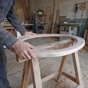 May include: A large, circular, unfinished light-coloured wooden frame with visible finger joints, resting on a wooden sawhorse. The frame features a smooth, rounded top edge and a flat inner surface, showcasing the natural grain of the raw wood material.