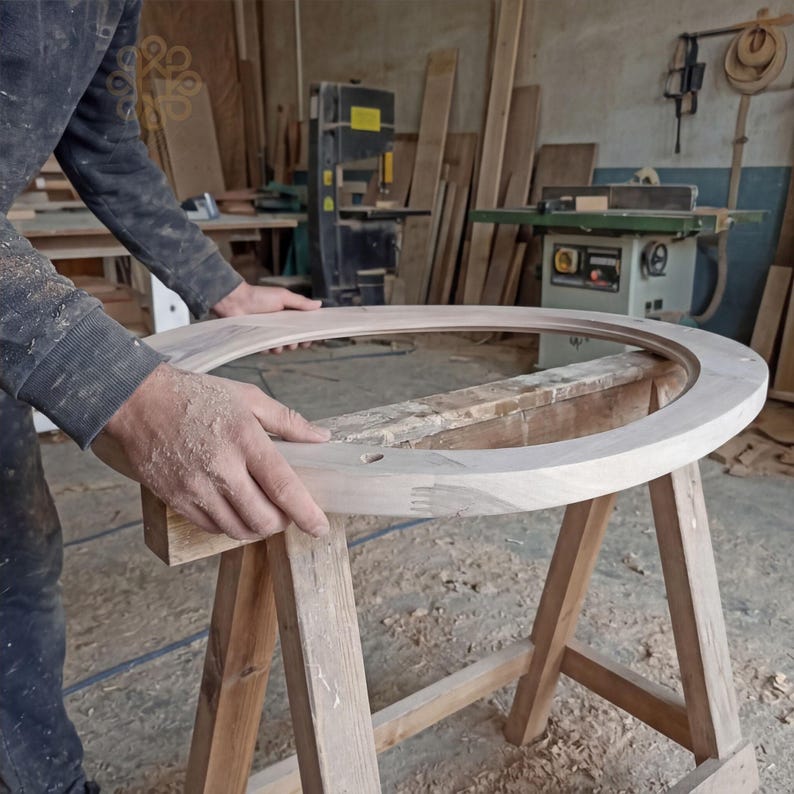 May include: A large, circular, unfinished light-coloured wooden frame with visible finger joints, resting on a wooden sawhorse. The frame features a smooth, rounded top edge and a flat inner surface, showcasing the natural grain of the raw wood material.