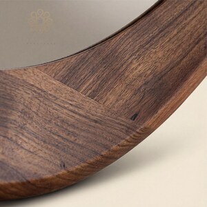May include: A circular mirror featuring a wide, polished dark brown wooden frame with a visible natural grain pattern. The frame is constructed from segments of wood joined together, and a small, faint logo is etched into the glass surface.
