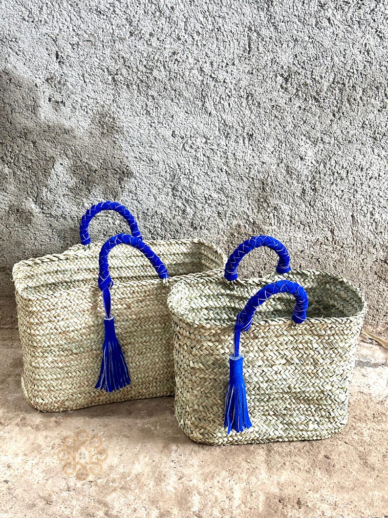 May include: Two woven straw tote bags with blue handles and tassels. The bags are rectangular with a textured weave pattern. The handles are a vibrant blue, and each bag has a matching blue tassel hanging from the handle.