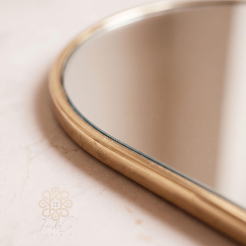 K&ouml;nnte beinhalten: Ein geschwungener Spiegel mit einem glatten, goldfarbenen Metallrahmen. Die reflektierende Oberfl&auml;che ist klar und glatt und zeigt eine subtile Spiegelung. Ein kleines, komplexes Logo mit dem Text Fes & Co. Home Decor ist unten links sichtbar.