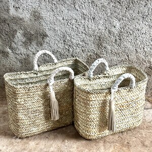 May include: Two woven straw tote bags with white braided handles and tassels. The bags are rectangular with a natural, light brown colour, set against a textured grey wall and a light brown surface.