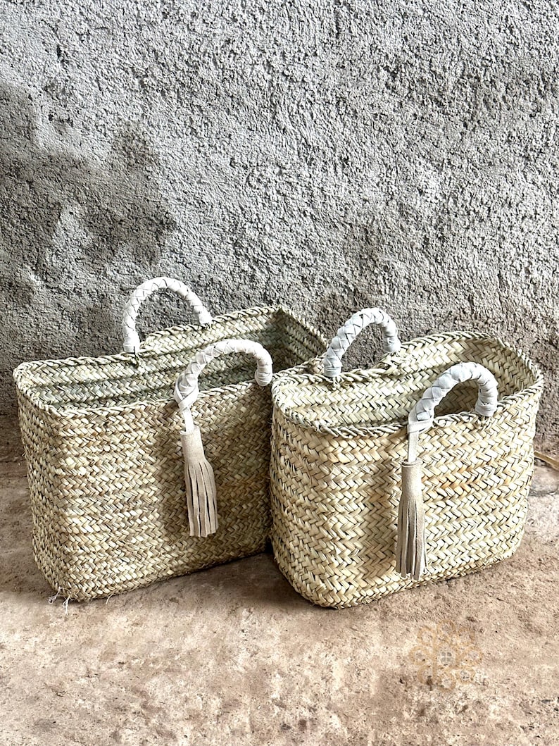 May include: Two woven straw tote bags with white braided handles and tassels. The bags are rectangular with a natural, light brown colour, set against a textured grey wall and a light brown surface.