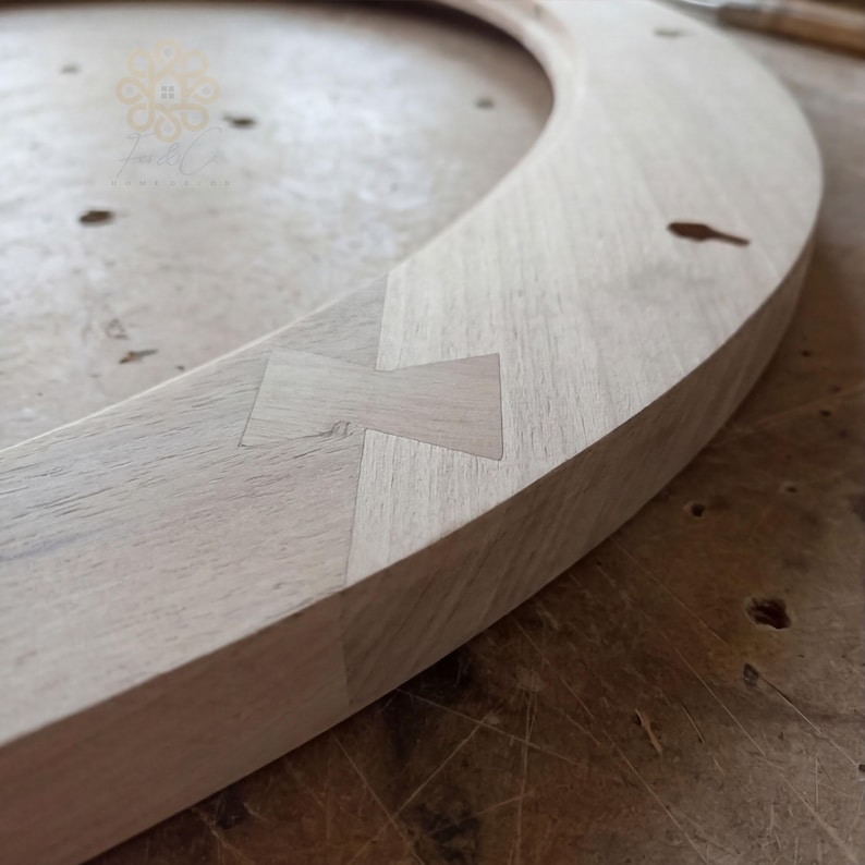 May include: A circular light-coloured wooden frame featuring a visible butterfly joint or bowtie inlay. The wood grain is clearly defined across the curved surface, which includes several small, dark, keyhole-shaped cutouts along the outer edge.