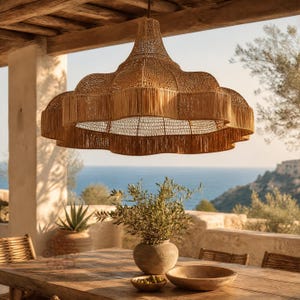 May include: A large, bell-shaped pendant light fixture crafted from woven natural tan fibers. The shade features a scalloped, multi-tiered edge with vertical fringe detailing, suspended from a single cord above a wooden outdoor dining table.