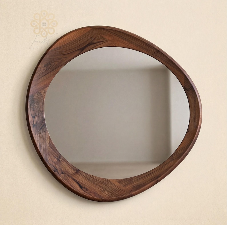 May include: A wall mirror with an irregular, organic, pebble-shaped frame made of dark brown wood with a visible grain pattern. The reflective glass surface is centred within the thick, asymmetrical wooden border, creating a smooth, rounded silhouette.