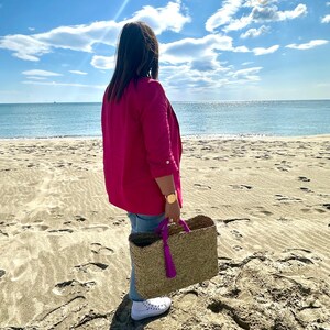 May include: A woman wearing a fuchsia blazer and holding a woven tote bag with purple handles and tassel, standing on a sandy beach. The tote bag is made of natural fibres. The background features a blue sky with clouds and the ocean.