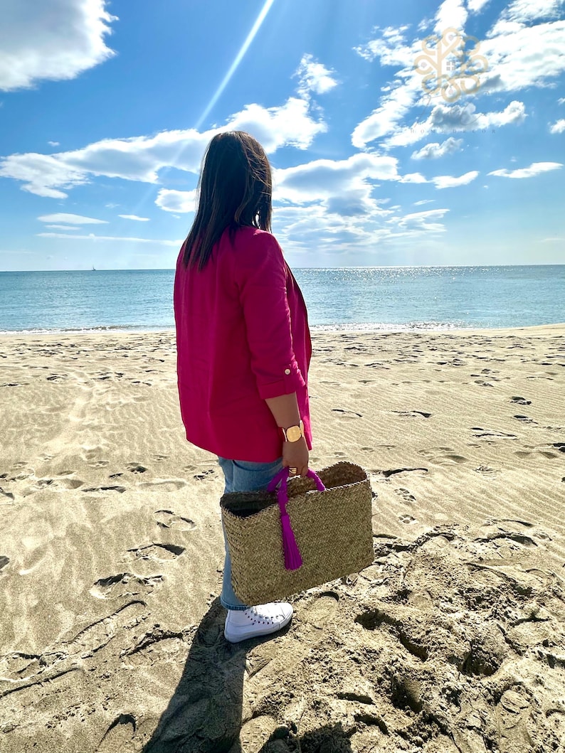 May include: A woman wearing a fuchsia blazer and holding a woven tote bag with purple handles and tassel, standing on a sandy beach. The tote bag is made of natural fibres. The background features a blue sky with clouds and the ocean.