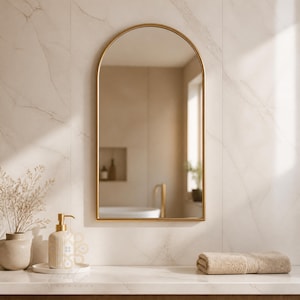 K&ouml;nnte beinhalten: Ein bogenf&ouml;rmiger Wandspiegel mit einem d&uuml;nnen goldenen Metallrahmen. Der Spiegel reflektiert ein Badezimmer mit einer wei&szlig;en Badewanne und einer Wandnische. Er ist an einer Wand mit hellen Marmorfliesen &uuml;ber einer wei&szlig;en Arbeitsplatte montiert.