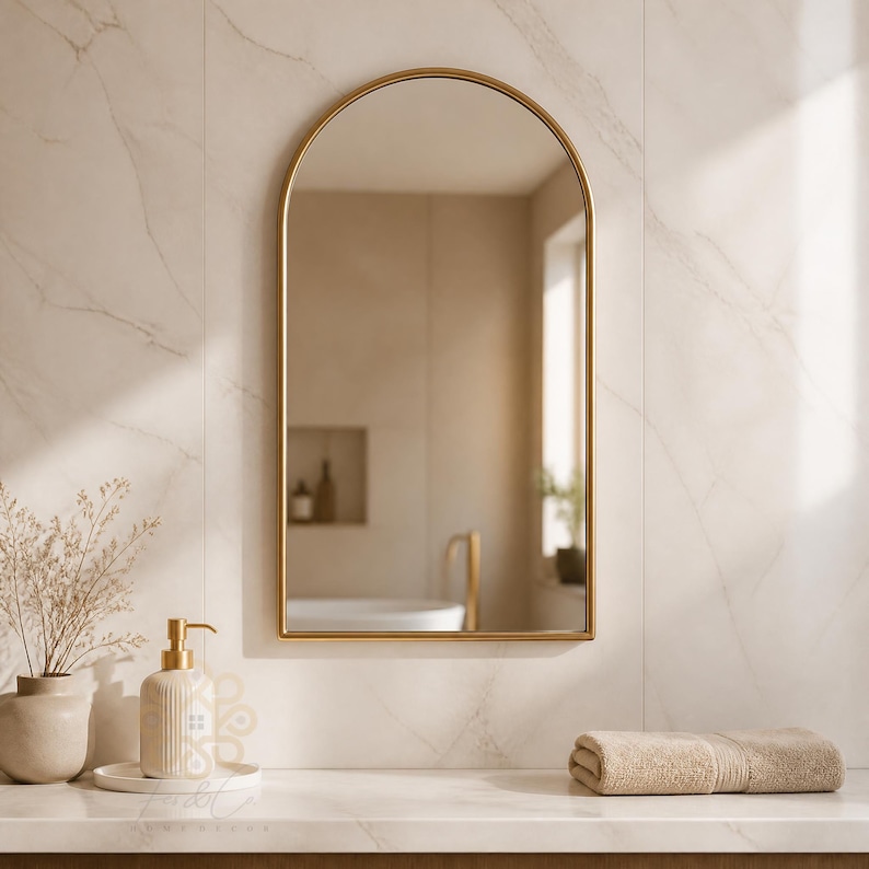 K&ouml;nnte beinhalten: Ein bogenf&ouml;rmiger Wandspiegel mit einem d&uuml;nnen goldenen Metallrahmen. Der Spiegel reflektiert ein Badezimmer mit einer wei&szlig;en Badewanne und einer Wandnische. Er ist an einer Wand mit hellen Marmorfliesen &uuml;ber einer wei&szlig;en Arbeitsplatte montiert.