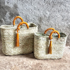 May include: Two woven straw tote bags. The bags have rectangular shapes with braided orange handles and tassels. The bags are different sizes, with the larger one on the left and the smaller one on the right. The bags are made of natural straw.