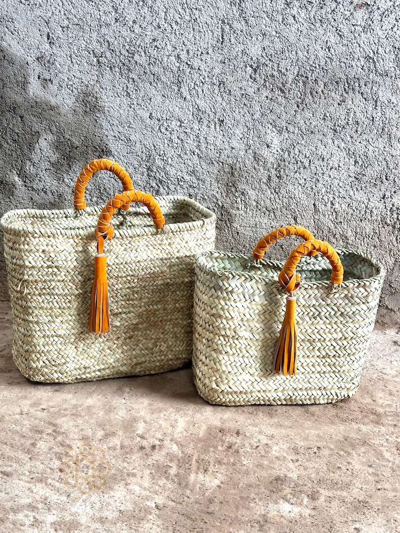 May include: Two woven straw tote bags. The bags have rectangular shapes with braided orange handles and tassels. The bags are different sizes, with the larger one on the left and the smaller one on the right. The bags are made of natural straw.