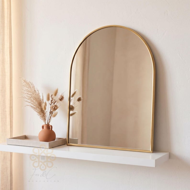 K&ouml;nnte beinhalten: Ein bogenf&ouml;rmiger Wandspiegel mit einem d&uuml;nnen, goldfarbenen Metallrahmen. Der Spiegel hat eine abgerundete Oberseite und gerade Seiten, die den umgebenden Raum reflektieren. Das Design ist schlicht und sauber und bietet eine klare Spiegelfl&auml;che.