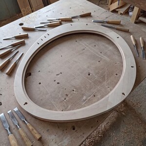 May include: A large, oval-shaped wooden frame with a smooth, light-coloured finish, featuring several small pre-drilled holes along its inner and outer edges, surrounded by an assortment of various metal woodworking chisels with wooden handles.