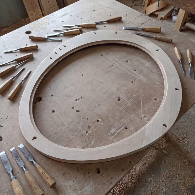 May include: A large, oval-shaped wooden frame with a smooth, light-coloured finish, featuring several small pre-drilled holes along its inner and outer edges, surrounded by an assortment of various metal woodworking chisels with wooden handles.
