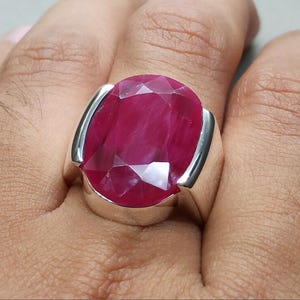 May include: A striking ruby ring featuring a large, oval-cut gemstone set in a silver band. The ruby displays a rich, deep red hue. The silver band has a minimalist design, complementing the gemstone.