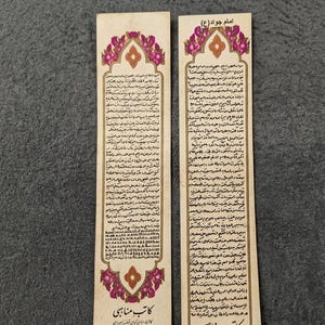 May include: Two rectangular paper bookmarks with ornate floral designs in pink and gold. Each bookmark features text in a script, with the title "Imam Javad (A)" at the top. The bottom of the bookmark has the text "Katib Manabi".