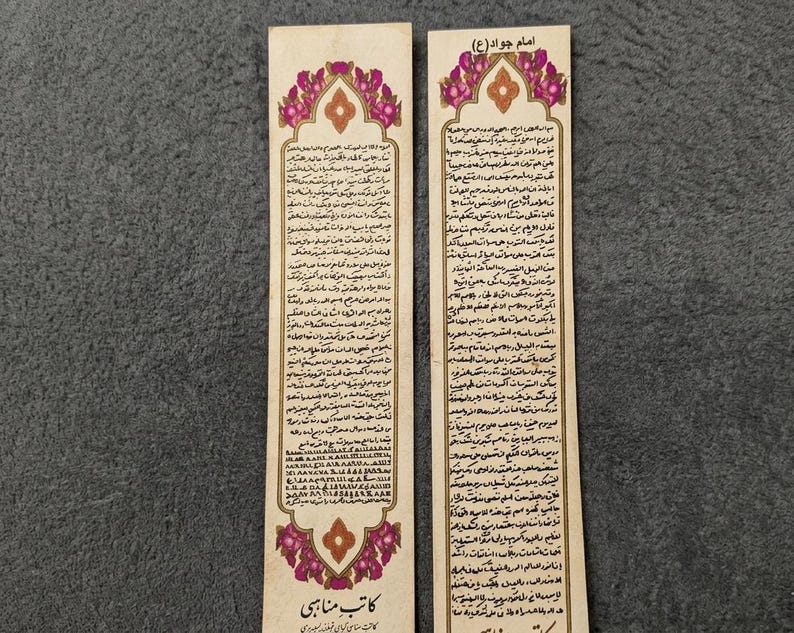 May include: Two rectangular paper bookmarks with ornate floral designs in pink and gold. Each bookmark features text in a script, with the title "Imam Javad (A)" at the top. The bottom of the bookmark has the text "Katib Manabi".