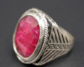 May include: A silver ring with a large, faceted, oval-shaped ruby gemstone. The ring has a detailed, openwork design on the sides and a rope-like border around the ruby. The ruby is a vibrant shade of red.