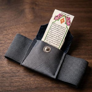 May include: A gray fabric wallet with a snap closure, holding a small card with text and floral accents. The wallet has an elastic band and is set against a wooden surface.