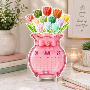 May include: A pink glass vase-shaped desk calendar for January 2027, featuring a bouquet of colorful tulips in red, orange, yellow, green, blue, and purple. The vase is adorned with a pink bow and a heart charm, displaying the month, year, and dates.