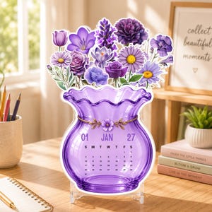 May include: A purple glass-style desk calendar for January 2027, featuring a scalloped rim and a gold floral accent. The calendar is topped with a vibrant arrangement of purple flowers, including roses, daisies, and lavender, all set on a clear stand.