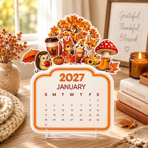 May include: A 2027 January calendar featuring a die-cut topper with autumn-themed illustrations including a hedgehog, squirrel, fox, orange tree, coffee cup, pumpkin, mushroom, and yellow rain boots, all displayed on a clear acrylic stand.