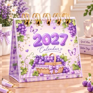 Op de afbeelding: Een bureaukalender voor 2027 met spiraalbinding, uitgevoerd in paars en wit met illustraties van druiventrossen, een rieten mand, vlinders en kleine hartjes. De omslag toont de tekst 2027 Calendar in een opvallend paars lettertype.