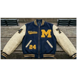 May include: A navy and cream varsity jacket featuring the word "Wolverines" and the letter "M" in yellow. The jacket has the number "24" and "4" on the sleeves. The jacket is zipped up and has a ribbed collar and cuffs.