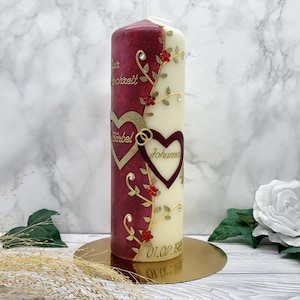 Könnte beinhalten: Eine dekorative Säulenkerze, vertikal geteilt in bordeauxfarbene und cremefarbene Abschnitte. Die bordeauxfarbene Seite zeigt goldene Schrift und Herz-Ausschnitte mit Namen. Blumengirlanden mit roten Akzenten und Strasssteinen verzieren die Kerze. Die Kerze steht auf einem goldenen Sockel.